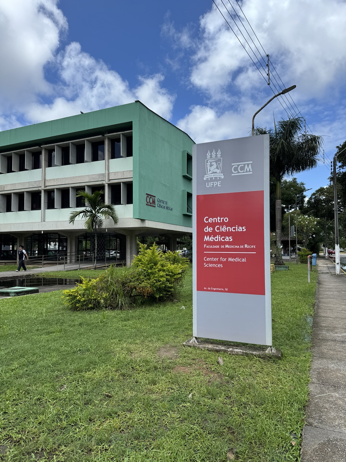 Foto real da fachada verde e da placa de identificação do Centro de Ciências Médicas da UFPE, em Recife